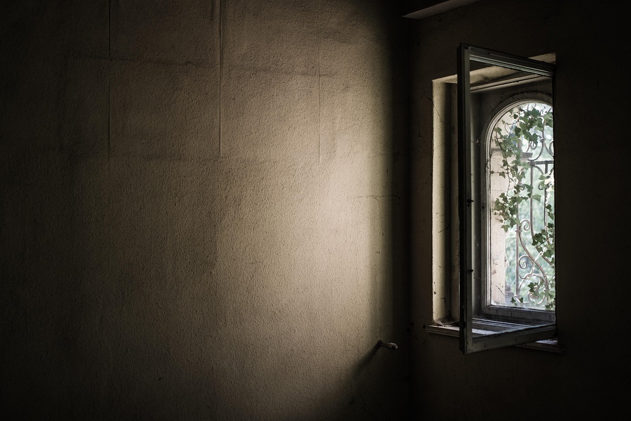 An open casement window admits a narrow patch of daylight into a sparse room with dark textured walls.