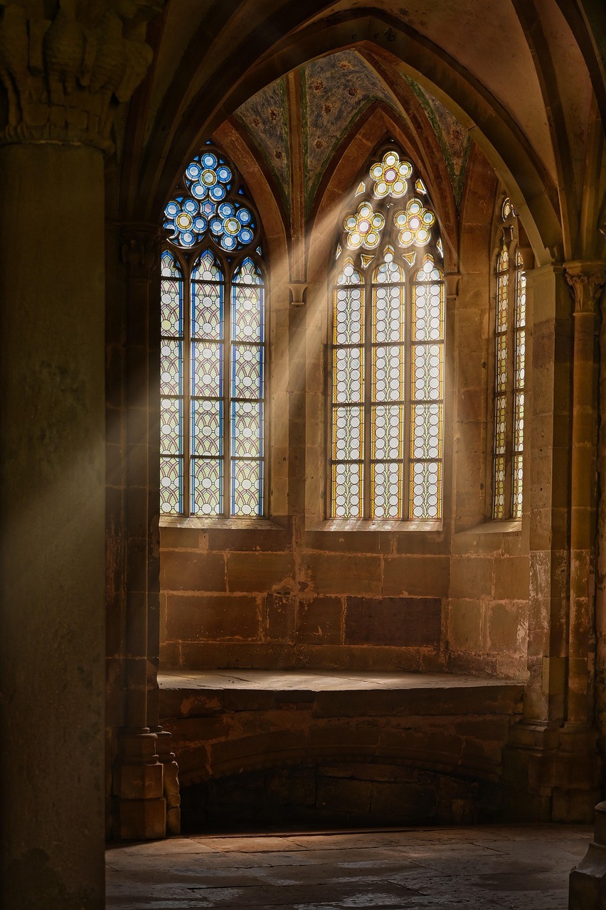 Tall pointed windows illuminate a stone church interior, with patterned glass and sunlit masonry surrounding a deep recess.