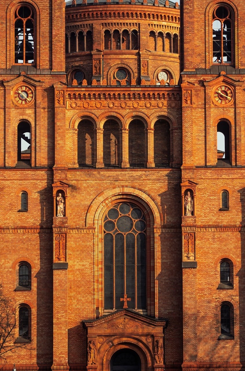A tall red-brick church façade features arched windows, circular details, towers and a large central recessed window.