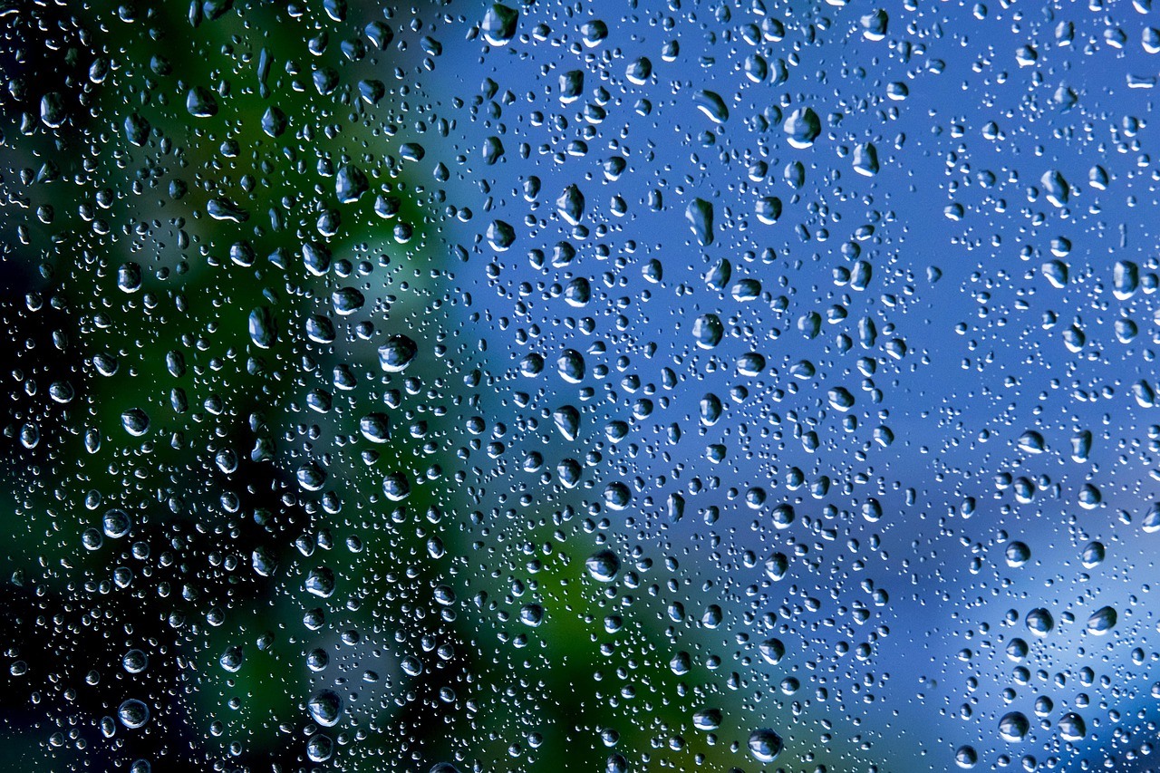 Numerous rain droplets cling to a pane, sharply visible against blurred blue sky and green foliage.